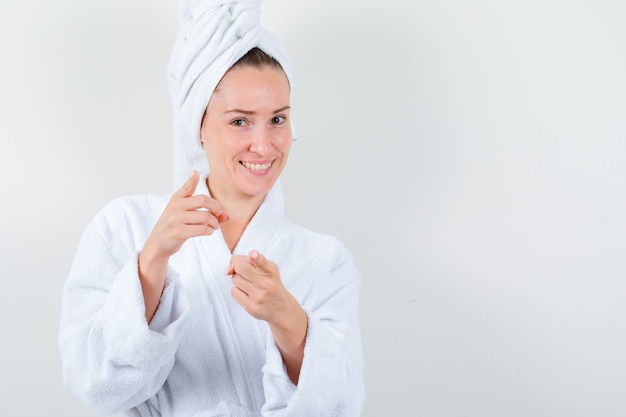 Young lady in white bathrobe, towel pointing at camera and looking joyful , front view.