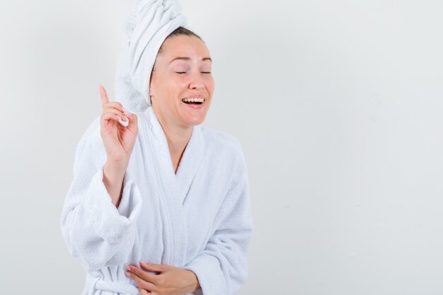 Young lady in white bathrobe, towel pointing up and looking happy , front view.