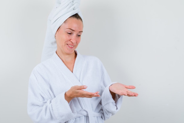 Young lady in white bathrobe, towel pretending to show something and looking joyful , front view.