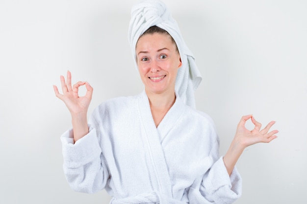 Young lady in white bathrobe, towel showing ok gesture and looking happy , front view.