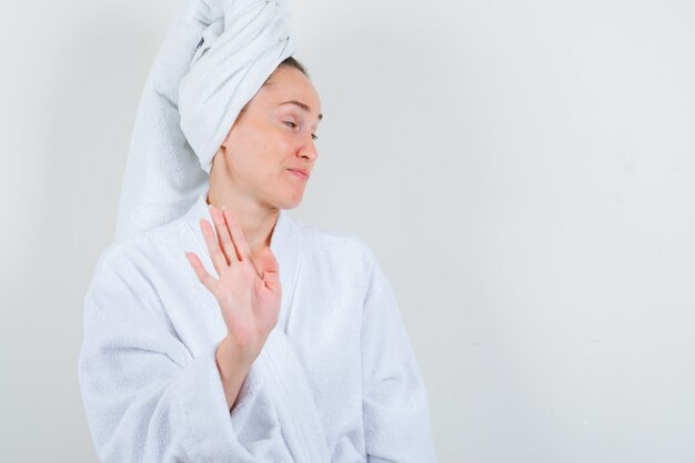 Young lady in white bathrobe, towel showing stop gesture and looking dissatisfied , front view.