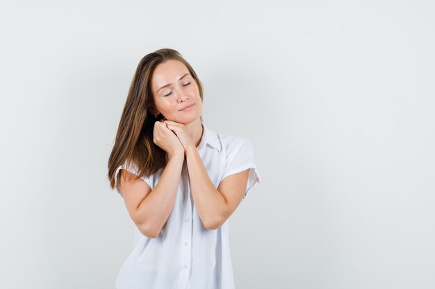 Young lady in white blouse closing her eyes while making pillow gesture and looking sleepy