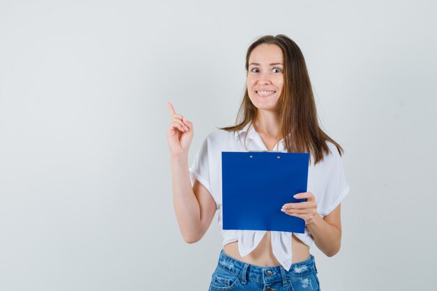 Young lady in white blouse holding clipboard while pointing away and looking hopeful , front view.