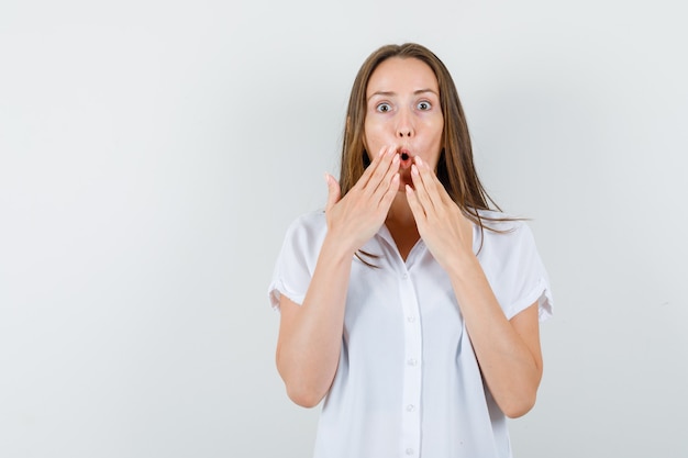 Young lady in white blouse holding hands on her chin and looking astonished