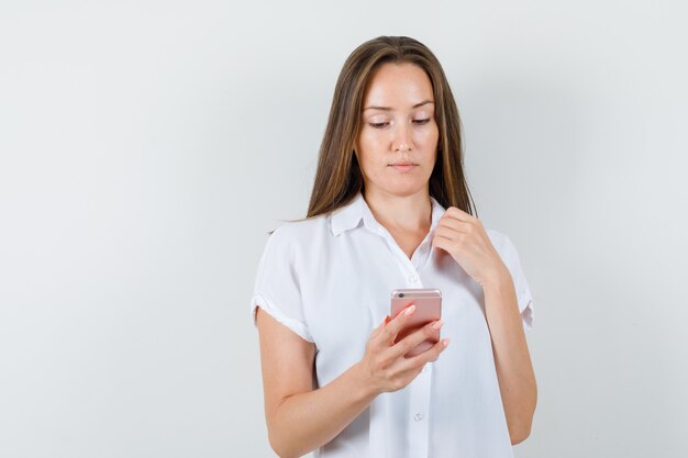 Молодая дама в белой блузке смотрит на телефон и смотрит сосредоточенно