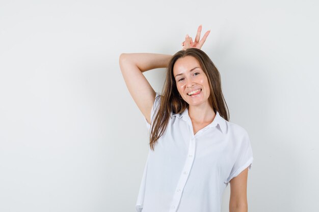 Девушка в белой блузке делает кроличий жест на голове и выглядит забавно