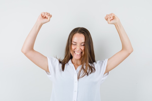 Young lady in white blouse raising her arms while showing fists and looking merry