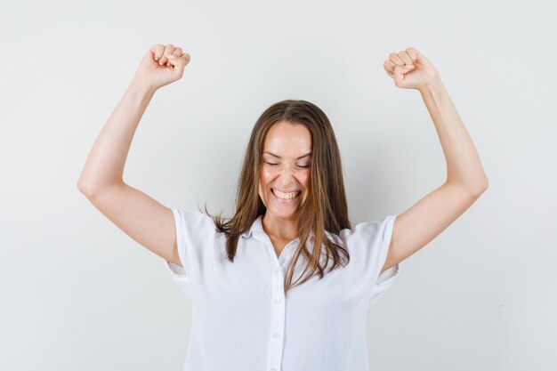 Young lady in white blouse raising her arms while showing fists and looking merry