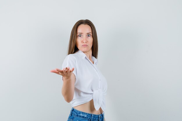Young lady in white blouse raising open palm and looking scared , front view.