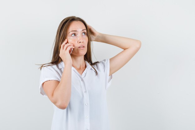 Young lady in white blouse talking on phone while holding hand on her head and looking thoughtful