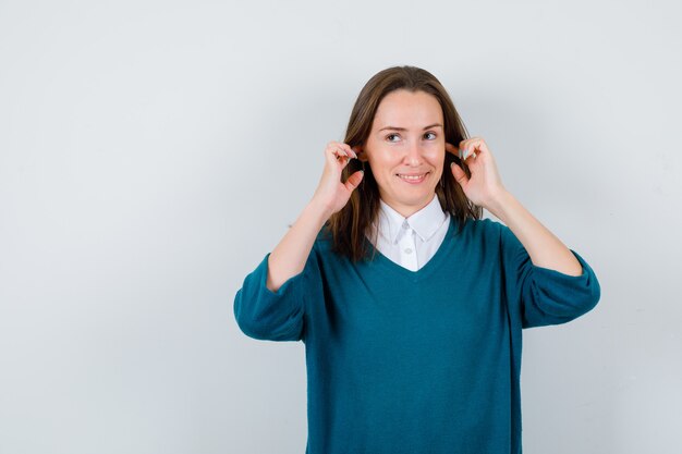 白いシャツを着た若い女性、指で耳を塞ぐセーター、目をそらし、陽気に見える、正面図。