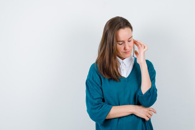 Young lady in white shirt, sweater touching cheek, keeping eyes shut and looking thoughtful , front view.