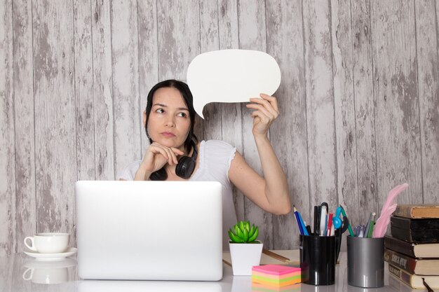 young lady in white t-shirt black earphones thinking holding white sign using grey laptop on table cup of coffee plant pens books on grey