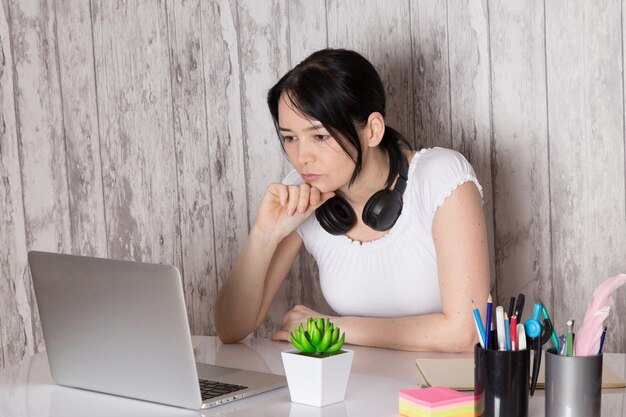 young lady in white t-shirt black earphones using grey laptop on table cup of coffee plant pens books on grey