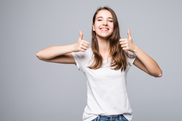 白いtシャツとジーパンの若い女性は白いスタジオ背景の前で歌う親指を示しています