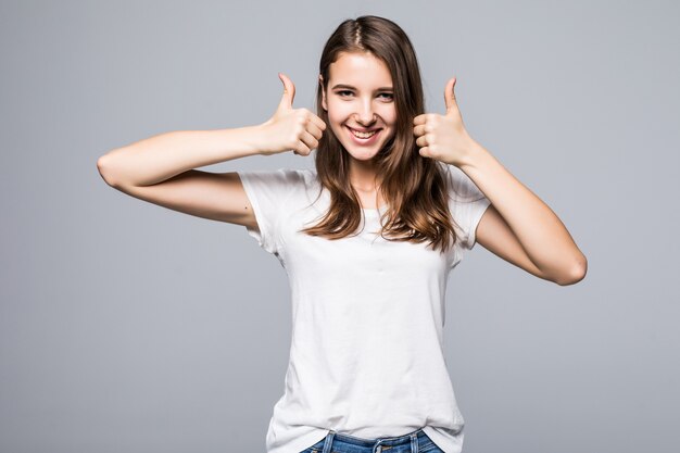 白いtシャツとジーパンの若い女性は白いスタジオ背景の前で歌う親指を示しています