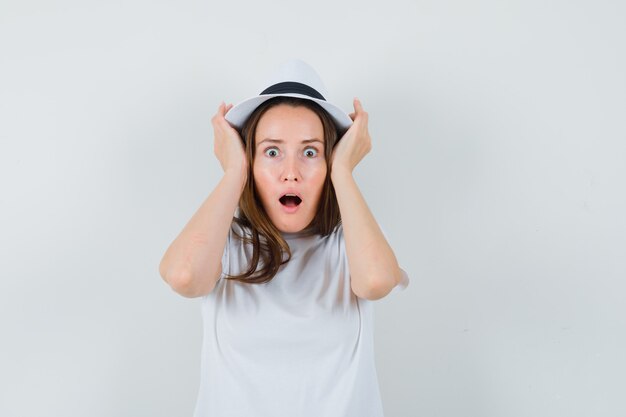 Young lady in white t-shirt hat holding hands to head and looking shocked  