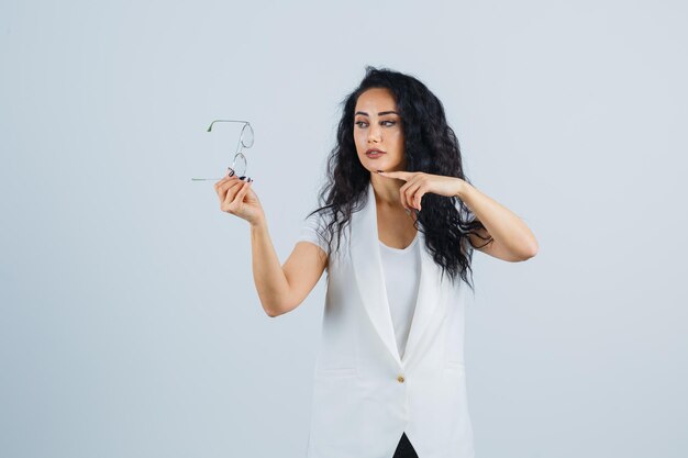 Young lady in white t-shirt, jacket pointing at glasses and looking confident , front view.