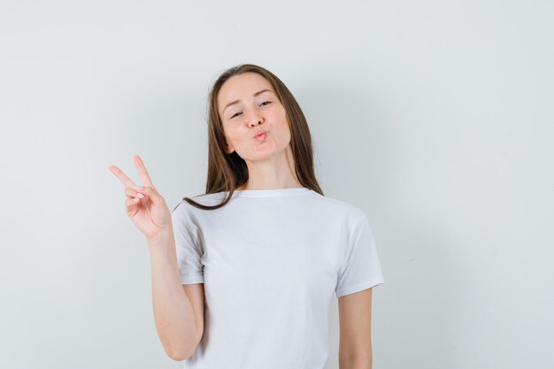 Молодая дама в белой футболке показывает жест победы, надувает губы и выглядит уверенно