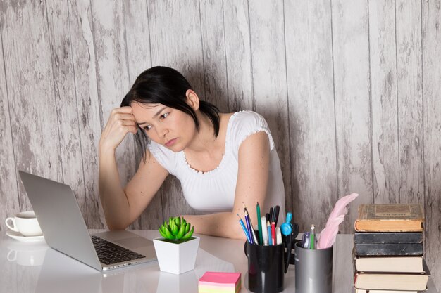 young lady in white t-shirt using grey laptop on table cup of coffee plant pens books on grey