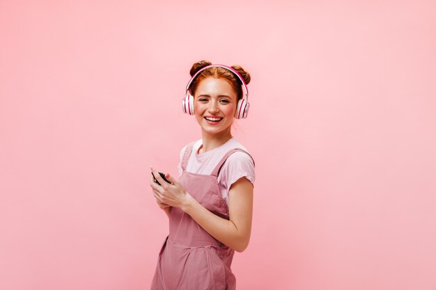 Девушка с булочками удивленно смотрит на экран телефона. Женщина в платье и белой футболке слушает музыку в наушниках на розовом фоне.