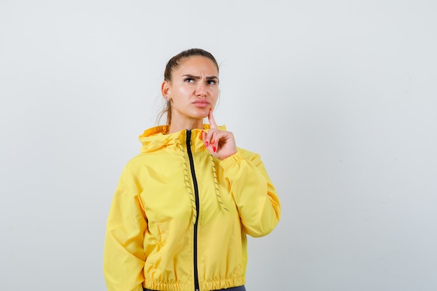 黄色いジャケットのあごに指を持ち、物思いにふける、正面図を探している若い女性。