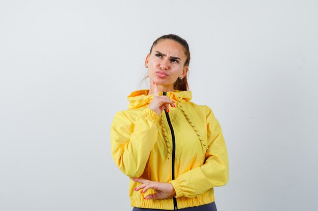 Young lady with finger on chin in yellow jacket and looking thoughtful. front view.