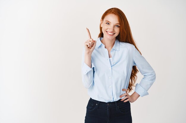 Young lady with ginger hair wearing blue shirt and pointing finger up, smiling as giving recommendation, demonstrating logo, white wall