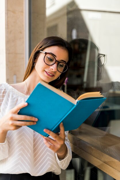 Young lady with glasses reading book leaning on window