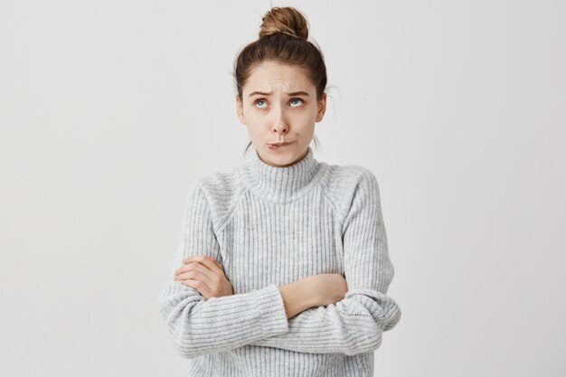 Young lady with hair in topknot standing with arms folded looking up. Capricious female model in grey dress biting lips thinking about her desire.  Daydream concept