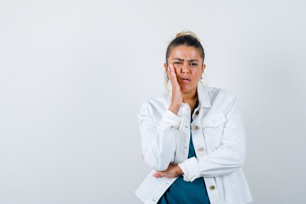 Young lady with hand on cheek in shirt, white jacket and looking serious , front view.