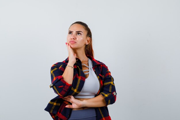 Young lady with hand on chin in top, plaid shirt and looking pensive. front view.