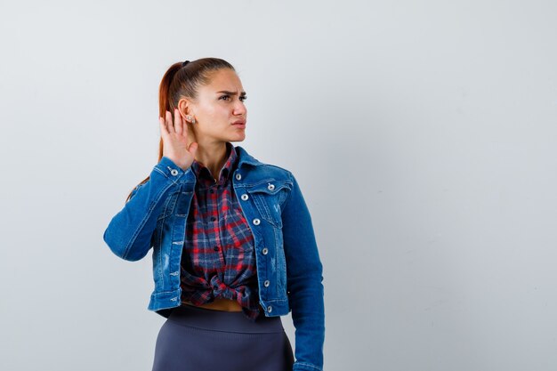 Young lady with hand behind ear in checkered shirt, denim jacket and looking careful. front view.