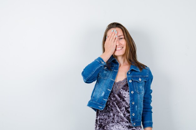 Young lady with hand on eye in blouse, denim jacket and looking cheerful , front view.