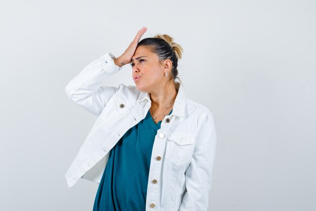 Young lady with hand on forehead in shirt, white jacket and looking forgetful. front view.