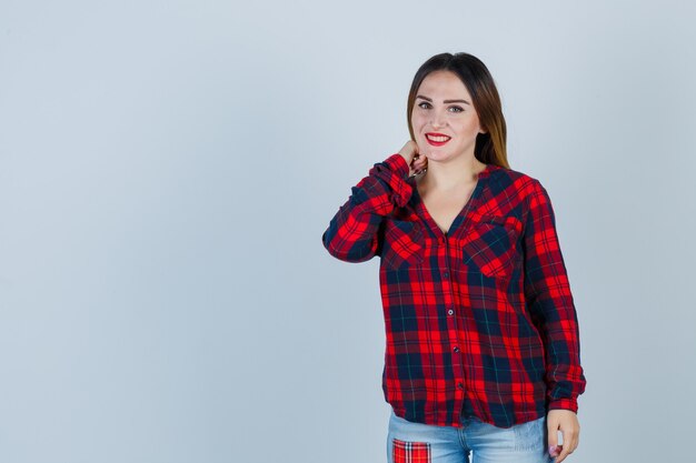 Young lady with hand on neck in checked shirt and looking ashamed , front view.