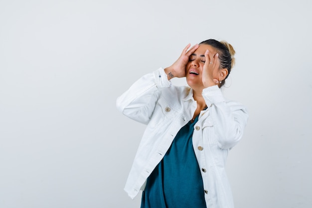 Young lady with hands on cheeks in shirt, white jacket and looking joyful , front view.