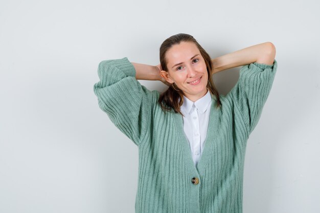 Young lady with hands behind head in shirt, cardigan and looking pleased , front view.