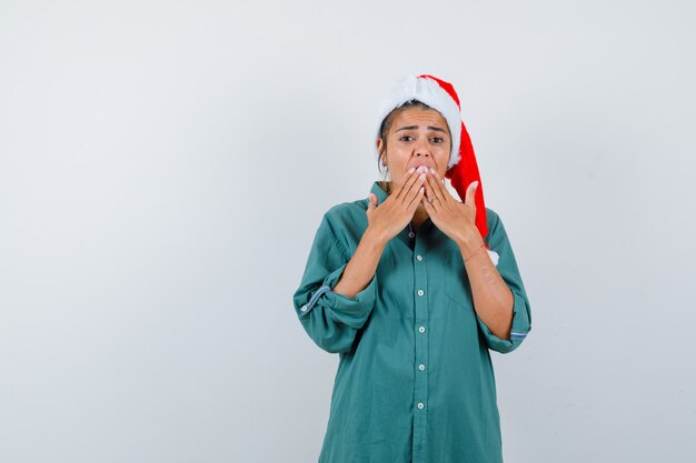 Молодая дама с руками на рту в рождественской шляпе, рубашке и испуганным видом. передний план.