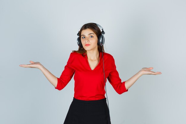 Young lady with headphones listening to music while making scales gesture in red blouse