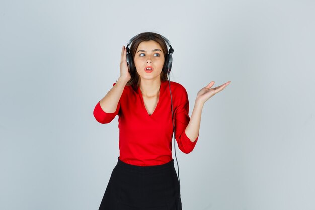 Молодая дама в наушниках слушает музыку, показывая что-то в красной блузке