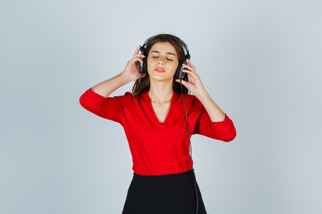 赤いブラウス、音楽を聴いて喜んでいるスカートのヘッドフォンを持つ若い女性