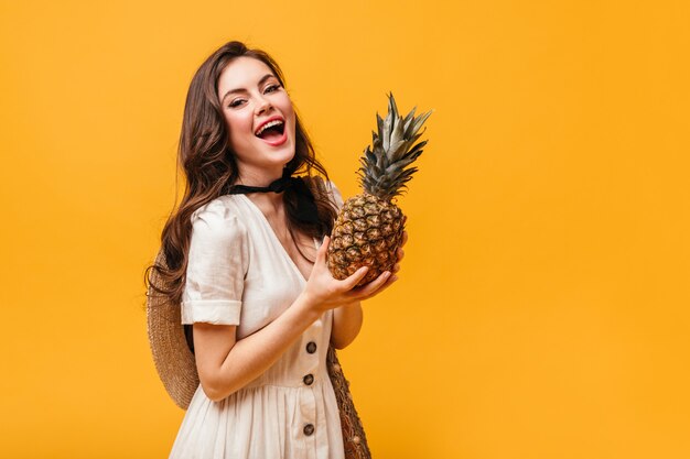 Young lady with nude makeup is holding pineapple. Woman in white dress laughs on orange background.