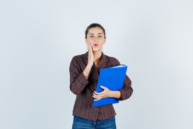 Young lady with palm on cheek in shirt, jeans and looking shocked, front view.