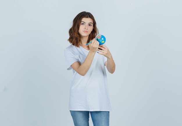 Young lady writing on globe with pencil while holding in t-shirt, jeans and looking focused. front view.