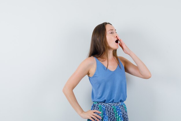 Young lady yawning with hand on mouth in blue blouse,skirt and looking tired , front view.