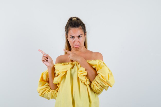 Young lady in yellow dress pointing at upper left corner and looking offended , front view.