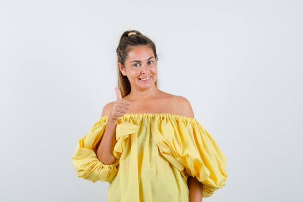 Young lady in yellow dress showing thumb up and looking merry , front view.