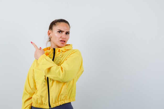 Девушка в желтой куртке, указывая на верхний левый угол и недовольно, вид спереди.