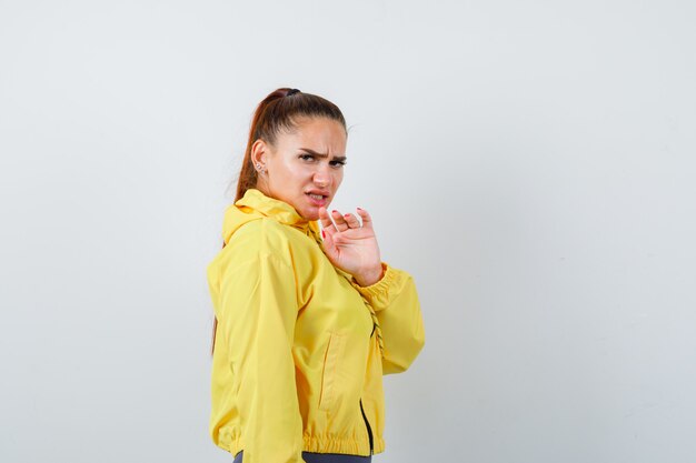 Молодая дама в желтой куртке поднимает руку, чтобы защитить себя и выглядит встревоженной, вид спереди.